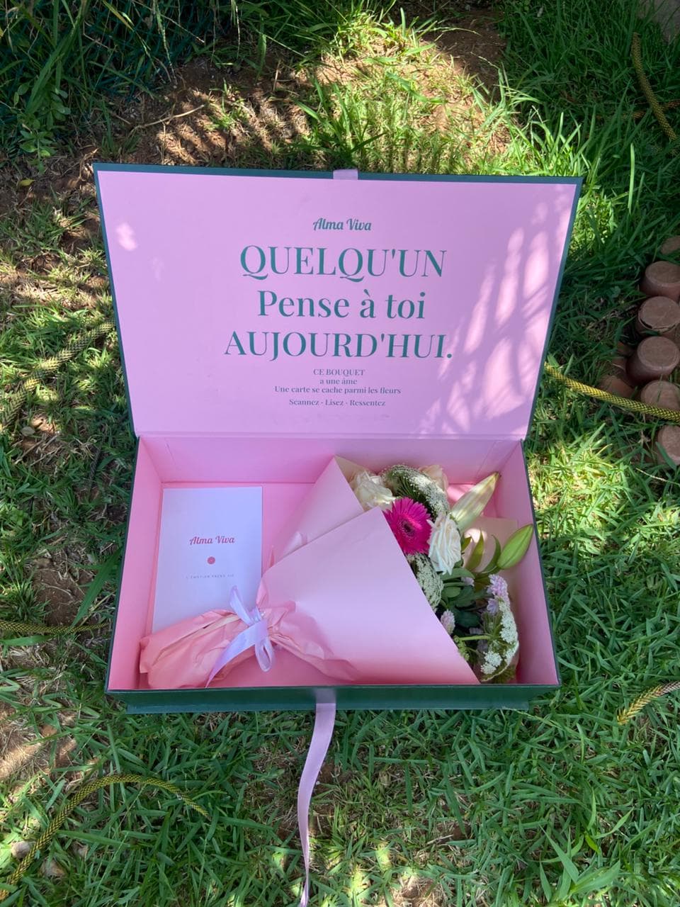 Boîte signature Alma Viva ouverte sur l'herbe, bouquet et carte à l'intérieur